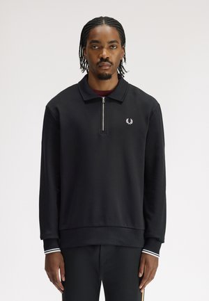 Mann mit geflochtenem Haar, der einen schwarzen, langärmeligen Sweatshirt mit Reißverschluss und weißem Lorbeer-Logo trägt und vor einfachem Hintergrund steht.