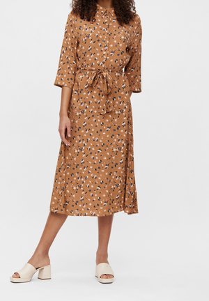 Mujer que lleva un vestido marrón de manga larga con botones, estampado floral y cinturón anudado en la cintura, combinado con sandalias crema de tacón cuadrado.