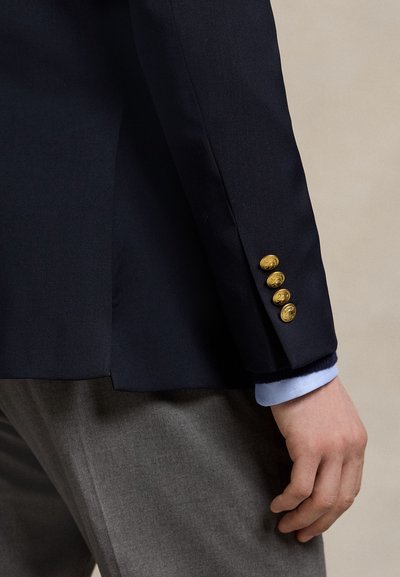 Højre arm af en person iført en navy blazer med fire gyldne knapper på ærmet og lys blå skjortemanchet, kombineret med grå bukser.
