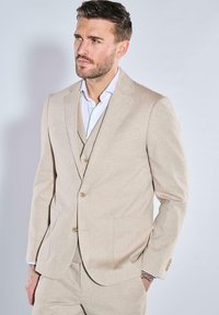 Hellbeige Anzug aus strukturiertem Stoff, mit zwei Knöpfen, Reverskragen und zwei Fronttaschen, getragen über einem weißen Hemd.