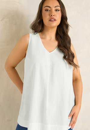 Femme aux longs cheveux bruns portant un haut blanc sans manches à col en V sur un fond beige uni.