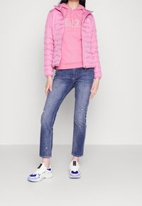 Giacca rosa trapuntata con cappuccio sopra un felpa abbinata con logo; jeans blu strappati; sneakers bianche e blu con lacci.