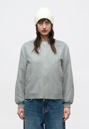 Grå bomberjakke med lynlås foran, ribbede manchetter og sidelommer. Kombineret med lyseblå, løsthængende cowboybukser og en lysegul hue.