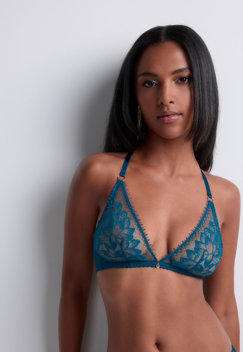 Bralette en dentelle teal avec motif floral, design triangulaire et fines bretelles. Présente des bordures décoratives et un petit détail en métal au centre.