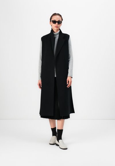 Melnā garā veste ar šallītes apkakli, apvienota ar pelēku turtleneck un salocītu melnu svārku. Pelēki loafers, melnas zeķes nodrošina piepildītu izskatu.