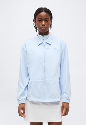 Frau mit kurzen geflochtenen Haaren trägt eine hellblaue Jacke mit Reißverschluss und weißen Rock, steht vor einem schlichten weißen Hintergrund.