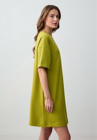 Abito t-shirt oversize verde lime, maniche corte, scollo rotondo, superficie liscia, lunghezza al ginocchio, tasche laterali, design semplice.