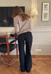 Femme aux longs cheveux, portant un pull beige et un jean noir, debout dans une pièce avec un sol en bois, une chaise, une lampe murale et une œuvre d'art encadrée représentant une plante.