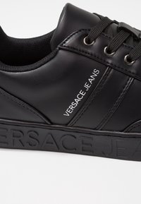 Baskets basses noires avec lacets noirs, "VERSACE JEANS" imprimé sur le côté et en relief sur la semelle.