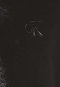 Tissu en maille noire avec un éclat subtil, présentant un logo en relief partiel "cK" sur le côté gauche. Texture lisse avec une légère variation.