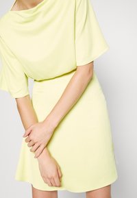 Vestido amarillo de manga corta con escote drapeado y silueta en A. Tela suave con un sutil brillo, que presenta costuras de manga suaves.