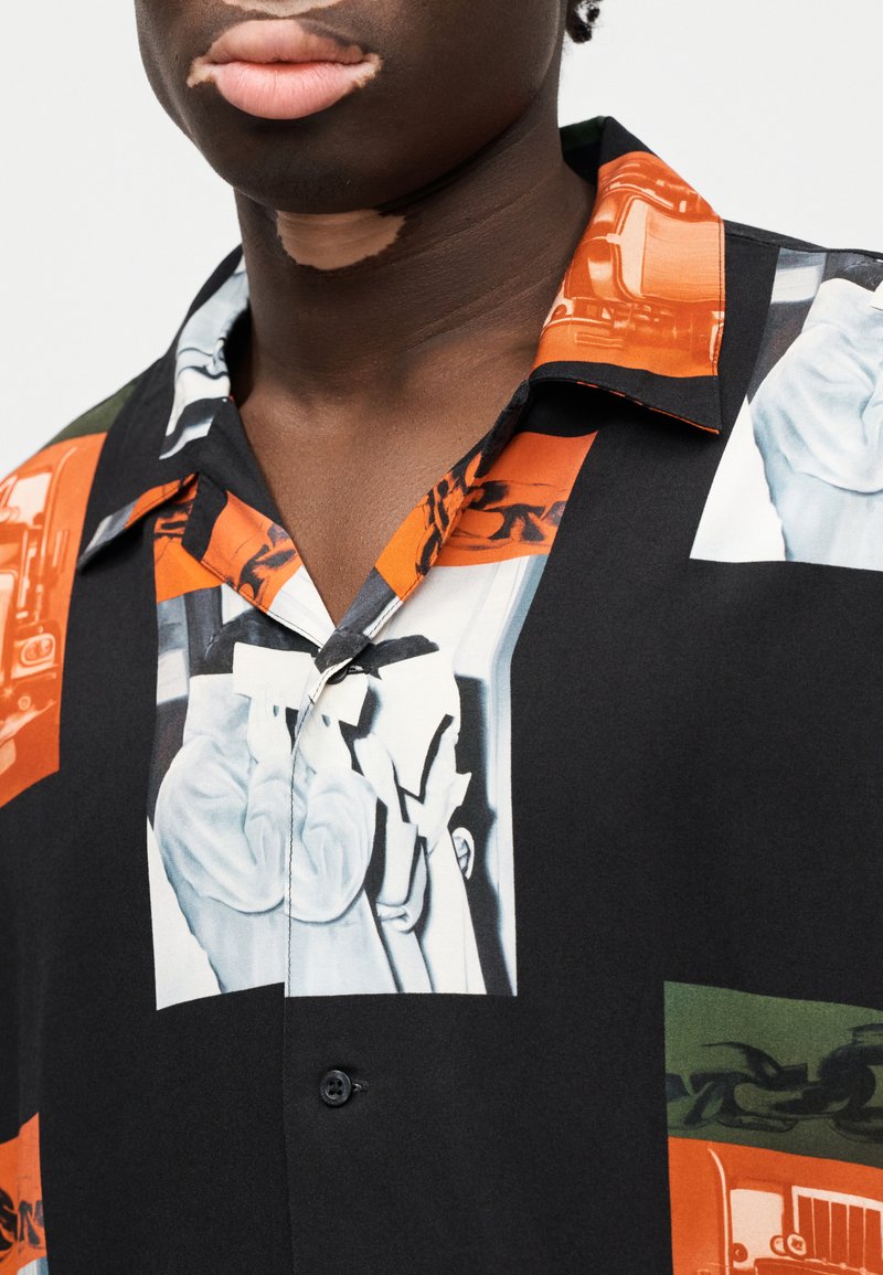 Camicia a maniche corte con stampa grafica audace in arancione, nero e verde. Il tessuto è liscio con colletto classico e chiusura con un solo bottone.