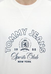 Άσπρο πουκάμισο με σκούρο μπλε κείμενο "Tommy Jeans Sports Club New York 1985" και έμβλημα με λιοντάρι και ονόματα πόλεων.