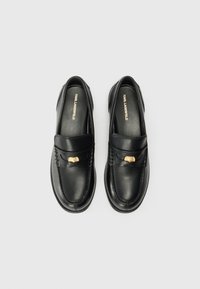 Schwarze Lederloafers mit glatter Oberfläche, ausgestattet mit einem goldenen Akzent am Riemen und genähten Details. Flacher Absatz und abgerunde Zehen.