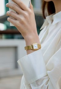 Wrist with gold rectangular watch and white dial, hand holding white cup near face, wearing white long-sleeve shirt.
