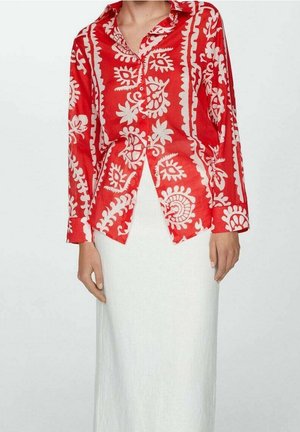 Kobieta ubrana w czerwoną bluzkę na guziki z białymi kwiatowymi wzorami i długą, prostą białą spódnicę na jednolitym tle.