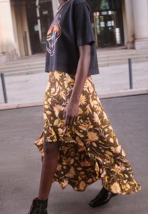Femme portant un t-shirt court graphique noir avec une jupe fluide jaune à motifs floraux et des bottines noires marchant en plein air.