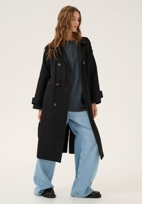 Černý dvouřadý trench coat s klopami a bočními kapsami, vrstvený přes modré tričko a široké modré džíny, doplněný tmavými botami.
