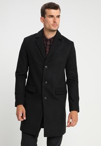 Cappotto lungo nero in misto lana con rever a torto, quattro bottoni, tasche frontali e una silhouette elegante, indossato sopra una camicia a quadri.