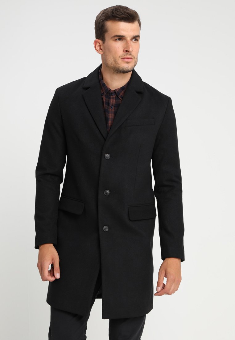 Cappotto lungo nero in misto lana con rever a torto, quattro bottoni, tasche frontali e una silhouette elegante, indossato sopra una camicia a quadri.