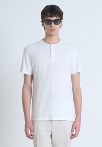 T-shirt bianco a maniche corte con scollatura rotonda, caratterizzato da una patta e una sottile texture. Indossato con occhiali da sole scuri su uno sfondo neutro.