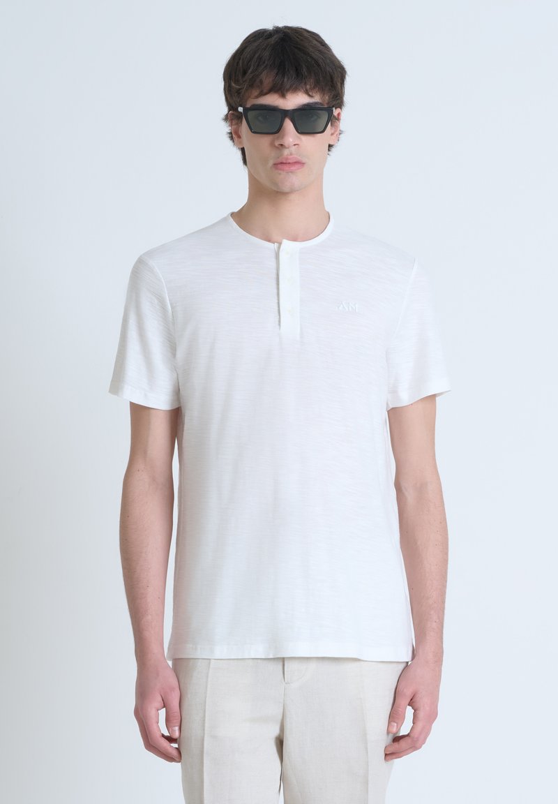 T-shirt bianco a maniche corte con scollatura rotonda, caratterizzato da una patta e una sottile texture. Indossato con occhiali da sole scuri su uno sfondo neutro.