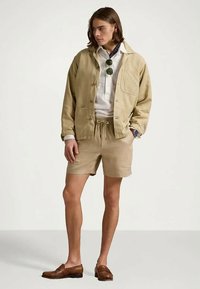 Jovem vestido com um casaco bege, calções a condizer, camisa branca, mocassins castanhos e óculos de sol pendurados na frente da camisa, de pé com uma mão no bolso.