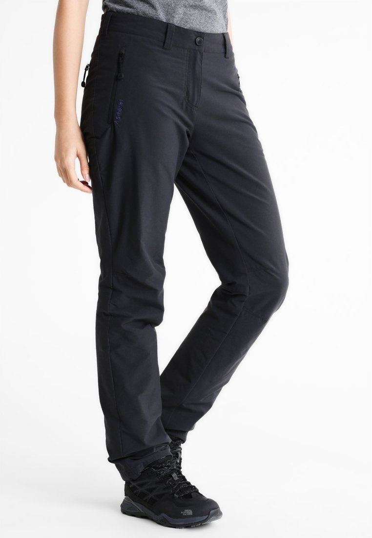 Personne portant un pantalon de plein air noir avec des poches latérales zippées et des chaussures de randonnée noires, debout avec une jambe pliée.