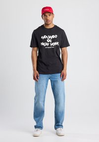 Black cotton t-shirt with "SOUNDS OF NEW YORK" text in white, paired with light blue denim jeans and a red cap. Sneakers complete the look.
