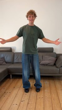 Zielona koszulka z krótkim rękawem z kieszonką na piersi, w połączeniu z luźnymi niebieskimi dżinsami i czarnymi butami, stojąca na drewnianej podłodze obok brązowej sofy.