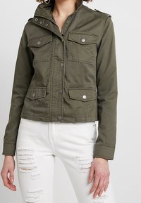 Olivově zelená zkrácená bunda z odolné látky, s předním zipem, dvěma náprsními kapsami s klopami a stříbrnými cvoky.