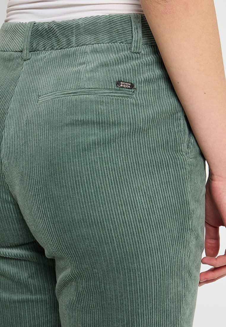 Grüne Cordhosen mit vertikaler Rippstruktur, ausgestattet mit einer Gesäßtasche und einem kleinen Logosticker auf der rechten Seite.