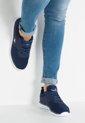 Person trägt hochgekrempelte blaue Jeans und dunkelblaue Kangaroos-Sneaker, ein Fuß lehnt an einer weißen Wand.