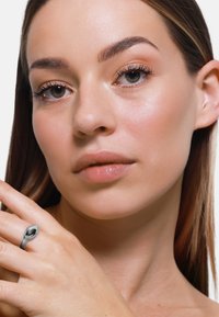 Anello in argento con un accento ovale nero, indossato su una mano su uno sfondo neutro; pelle liscia con trucco naturale, tratti del viso morbidi visibili.