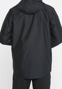 Czarna wodoodporna kurtka z kapturem, o gładkiej fakturze i luźnym kroju. Dół kurtki sięga poniżej talii, bez widocznych elementów metalowych ani wzorów.