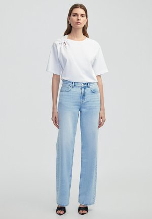 Witte katoenen T-shirt met een knoopdetail op de linkerschouder, gecombineerd met lichtblauwe high-waisted straight-leg jeans. Zwarte open-toed schoenen.
