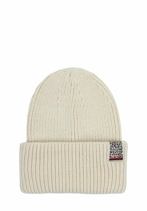 Knit beanie in cream color with a folded brim. Features a leopard print tag with red and blue accents at the side. Soft texture.