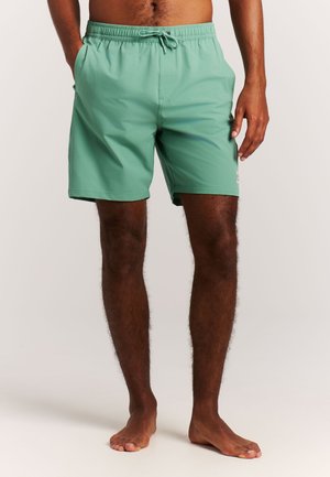 Uomo che indossa pantaloncini da bagno verde chiaro semplici con coulisse in vita, in piedi a piedi nudi su uno sfondo neutro.