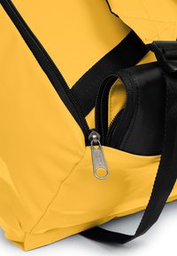 Mochila de tela amarilla con acentos negros, un bolsillo con cremallera y costuras duraderas. La bolsa tiene forma rectangular y correas ajustables.