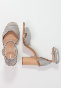 Sandali con tacco in pelle scamosciata grigia con punta aperta, cinturini incrociati e tacco a blocco in legno chiaro. Cinturino regolabile alla caviglia per una vestibilità perfetta.