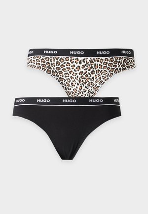 Žena s dlhými tmavými vlasmi, ktorá má na sebe leopardí podprsenku a spodnú bielizeň s čiernymi pásmi značky Hugo, stojí pred jednofarebným pozadím.