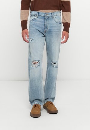 Man in lichtblauwe gescheurde jeans en bruine schoenen, gefotografeerd van het midden van de torso tot de voeten tegen een witte achtergrond.