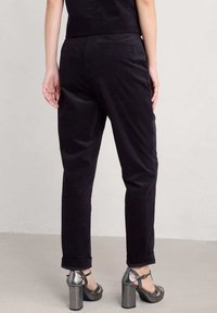 Črne korduro hlače z oprijetim krojem in zvitimi manšetami. Model nosi kovinske sandale s peto, kar razkriva zadnje žepke.