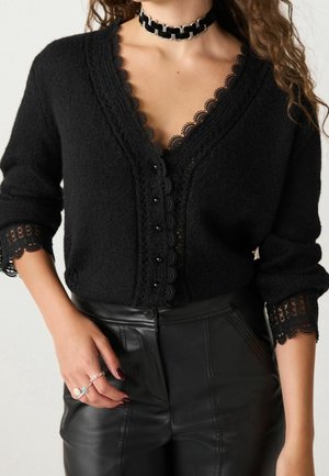 Cardigan noir bordé de dentelle porté sur un haut en dentelle, pantalon en cuir noir taille haute, bagues argentées et collier ras-du-cou en chaîne noire épaisse sur le mannequin.