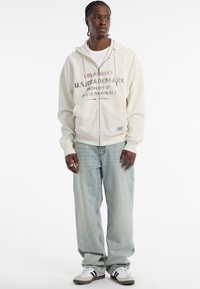 Cremfarbener Reißverschluss-Hoodie mit Frontgrafik, kombiniert mit hellblauen lässigen Jeans und weißen Sneakers mit schwarzen Streifen.