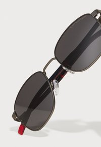 Occhiali da sole con montatura in metallo grigio opaco, lenti scure e dettagli rossi sulle aste. Presentano cuscinetti nasali trasparenti e un design piatto.