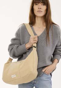 Sacoche beige fabriquée en tissu durable avec une texture douce, comportant une poche zippée à l'avant et une large bandoulière.