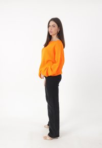 Pomarańczowy sweter z teksturą i długimi rękawami, zestawiony z czarnymi spodniami o wysokim stanie i beżowymi sandałami, na białym tle.