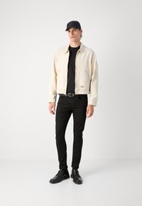 Chaqueta de mezclilla beige clara con cremallera, llevada sobre una camiseta negra. Jeans negros ajustados y zapatos negros de cordones completan el conjunto.