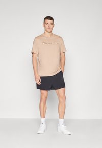 Beige Baumwoll-T-Shirt mit "Champion"-Logo in geschwungener Schrift, kombiniert mit navyblauen Shorts und weißen Turnschuhen. Model in stehender Pose.
